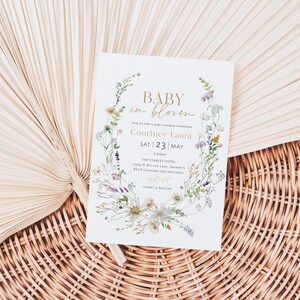 May include: A white baby shower invitation with a floral wreath design. The text reads "Baby in Bloom" and includes the name "Courtney Laura" and the date "Sat 23 | May".