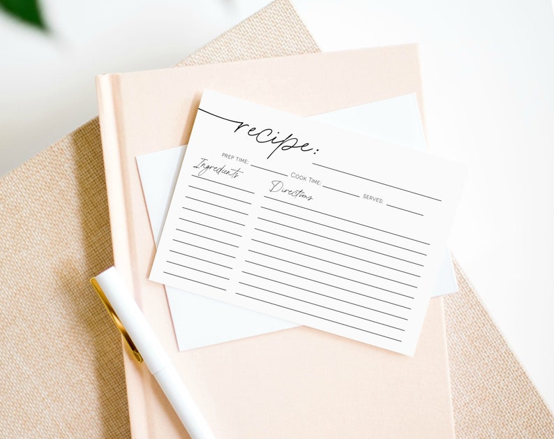 May include: A stack of recipe cards with the word "recipe" in cursive script at the top. The cards have lines for ingredients, directions, prep time, cook time, and servings. A white pen with a gold clip rests on a pink notebook.
