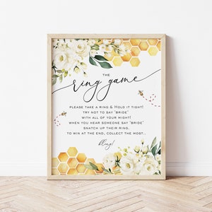 May include: A printable bridal shower game card with a white background and a honeycomb pattern. The text reads "The Ring Game" and includes instructions for the game. The card is decorated with white flowers and green leaves.
