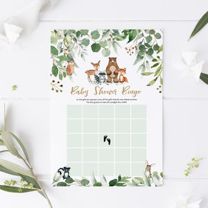 May include: A green and gold baby shower bingo card with a woodland animal theme. The card has a grid of squares with images of a bear, owl, deer, raccoon, fox, skunk, bunny, and footprints. The text "Baby Shower Bingo" is at the top of the card.
