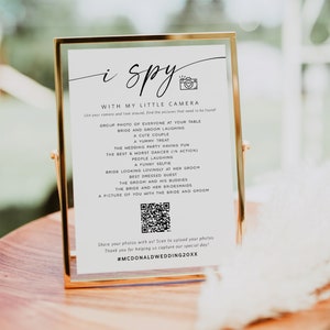 May include: A white sign with a gold frame, with the text "i spy with my little camera" and a list of items to find in wedding photos. The sign also includes a QR code and the hashtag #MCDONALDWEDDING20XX.