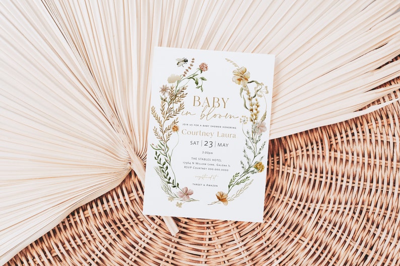 May include: A white baby shower invitation with a floral wreath border. The text reads "Baby in Bloom" and includes the name "Courtney Laura" and the date "Sat 23 | May".