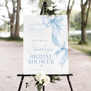 Op de afbeelding: Een wit bord op een zwart schildersezel met de tekst "Something Blue Before Saying I Do" en "Welcome to Charley's Bridal Shower." Een lichtblauw lintontwerp siert het bord. Het bord zegt ook "46 dagen totdat ze I Do zegt!"