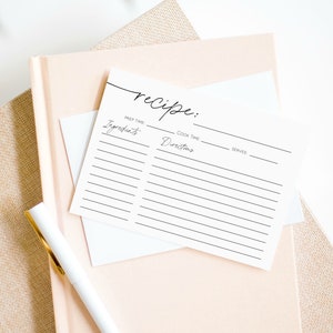 May include: A stack of recipe cards with the word "recipe" in cursive script at the top. The cards have lines for ingredients, directions, prep time, cook time, and servings. A white pen with a gold clip rests on a pink notebook.