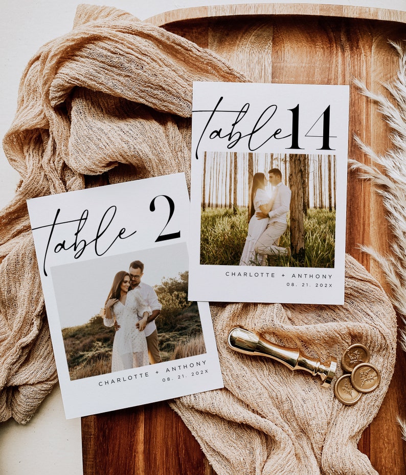May include: Two white table number cards with black calligraphy. The cards feature a couple's photo and the text "Table 2" and "Table 14" with the names Charlotte and Anthony and the date 08. 21. 202x.