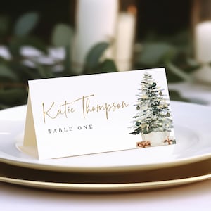 Peut inclure: Une carte de table blanche avec une inscription dorée qui indique "Katie Thompson" et "Table One". Une illustration aquarelle d'un sapin de Noël avec des ornements et des cadeaux se trouve sur le côté droit de la carte.