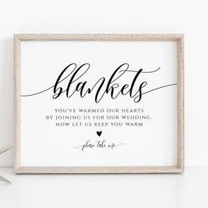 Puede incluir: Un cartel blanco con texto negro que dice "blankets" en una fuente cursiva. El texto debajo dice "You've warmed our hearts by joining us for our wedding. Now let us keep you warm."  El cartel también incluye un pequeño corazón negro y el texto "please take one" en una fuente cursiva.