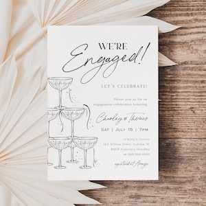 Könnte beinhalten: Eine Verlobungsankündigungskarte mit dem Schriftzug "We're Engaged!" in eleganter Schrift. Die Karte zeigt einen Stapel Champagnergläser und Veranstaltungsdetails, einschließlich der Namen Charley & Thomas, Datum, Uhrzeit und Ort.