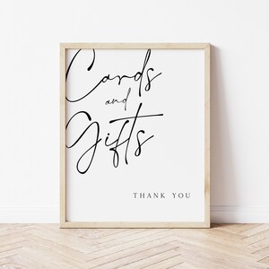 以下が含まれることがあります： 黒い枠のある白い看板に、筆記体で「Cards and Gifts」と書かれています。看板には、より小さなフォントで「Thank You」という文字も書かれています。
