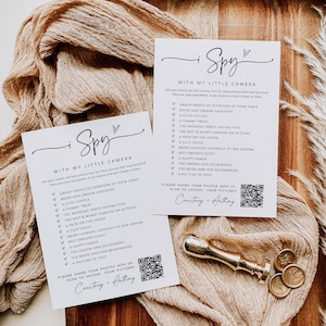 May include: A white card with black text that says "I Spy with my little camera" and lists a series of things to find in a wedding photo. The card also includes a QR code and the text "Please share your photos with us. Scan to upload your pictures. Courtney + Anthony."