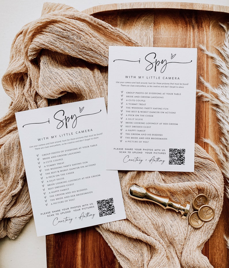 May include: A white card with black text that says "I Spy with my little camera" and lists a series of things to find in a wedding photo. The card also includes a QR code and the text "Please share your photos with us. Scan to upload your pictures. Courtney + Anthony."