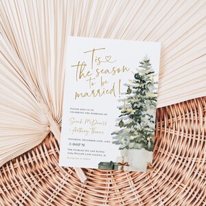 May include: A white wedding invitation with gold lettering that reads "Tis the season to be married!"  The invitation features a watercolor illustration of a Christmas tree with ornaments and presents. The text also includes the names "Sarah McDonald" and "Anthony Thomas" and the date "December 10th, 20XX".