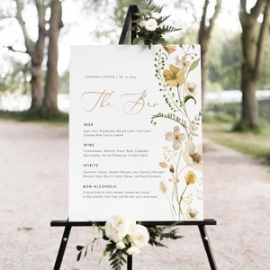 May include: A white sign with a floral design and gold lettering lists the bar menu for a wedding. The sign reads "The Bar" and lists beer, wine, spirits, and non-alcoholic options. The sign is on a black easel.