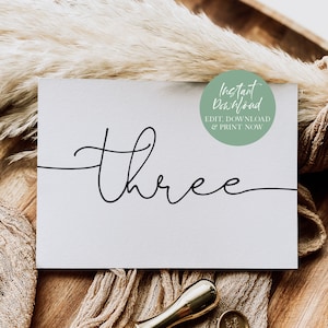May include: A white card with the word "three" written in black cursive script. The card is on a wooden surface with a brown fabric and a green circle with the text "Instant Download EDIT, DOWNLOAD & PRINT NOW".