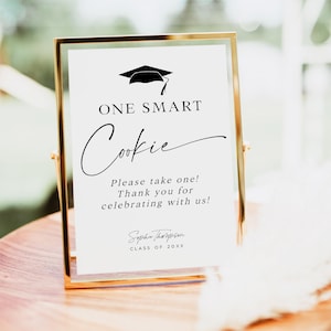 May include: A gold-framed sign with a white background and black text that reads "ONE SMART Cookie Please take one! Thank you for celebrating with us! Sophie Thompson CLASS OF 20XX". A black graduation cap icon is at the top of the sign.