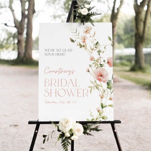 May include: A white sign with a floral design and the text "We're so glad your here! Courtney's Bridal Shower Saturday, July 15th 2021".