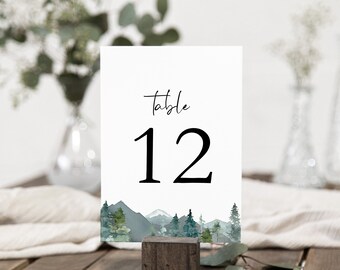 Op de afbeelding: Witte kaart met zwarte tekst die "Tafel 12" aangeeft en een aquarel-illustratie van een bergketen met bomen aan de onderkant.
