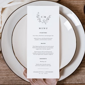 Könnte beinhalten: Eine Hochzeitsmenükarte mit einem schlichten Blumendesign und den Initialen "CM" in einem Kranz. Die Speisekarte listet Vorspeisen, Hauptgerichte und Desserts für die Hochzeit von Charlotte und Anthony am 10.01.2014 auf.