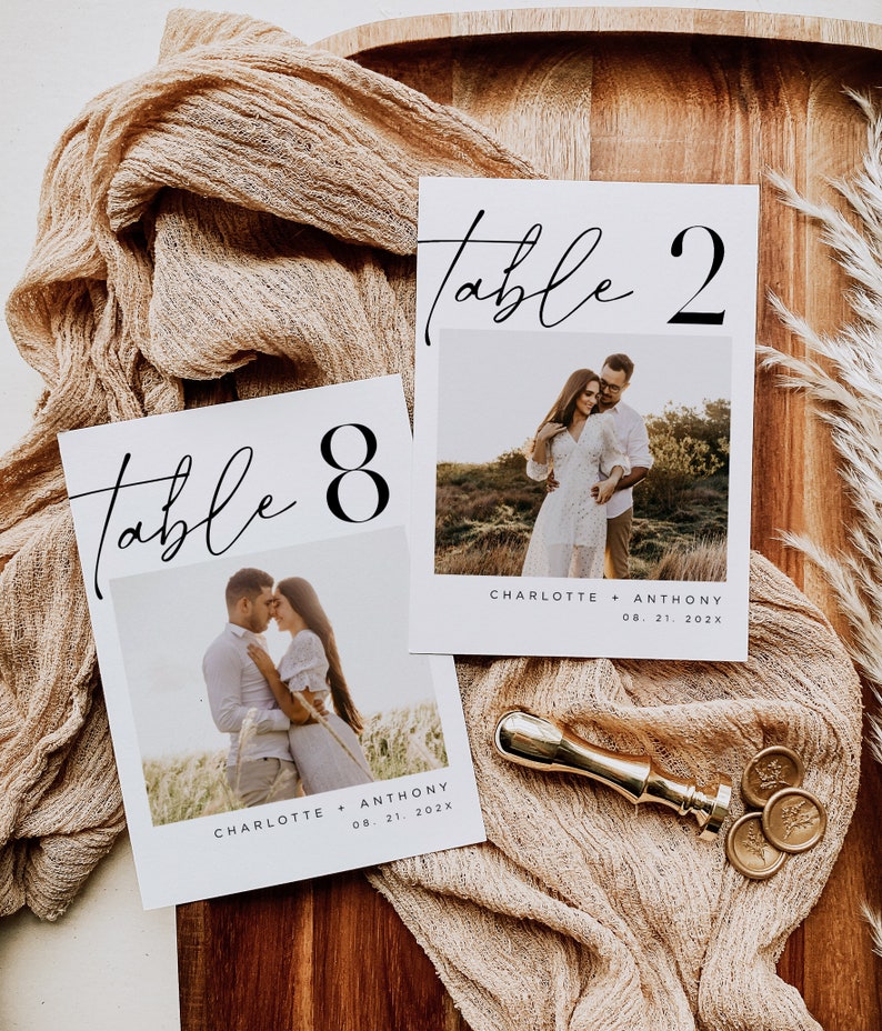 May include: Two white table number cards with black calligraphy and numbers. Each card features a photo of a couple in a field. The text on the cards reads "Table 8" and "Table 2". The cards also include the names "Charlotte + Anthony" and the date "08. 21. 202x".