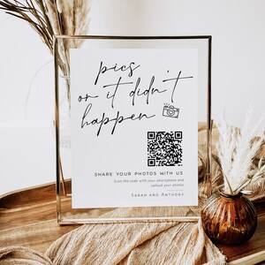 May include: A white sign in a gold frame with the text "pics or it didn't happen" and a QR code. The sign is on a wooden table with a vase of dried flowers.