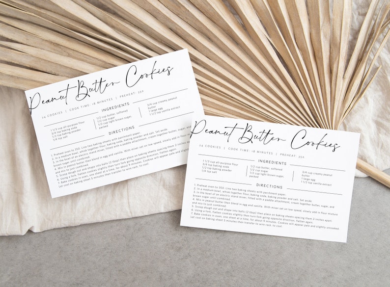 May include: Two printable recipe cards for peanut butter cookies. The cards are white with black text and include the ingredients and directions for making the cookies. The cards are laid out on a light brown surface.