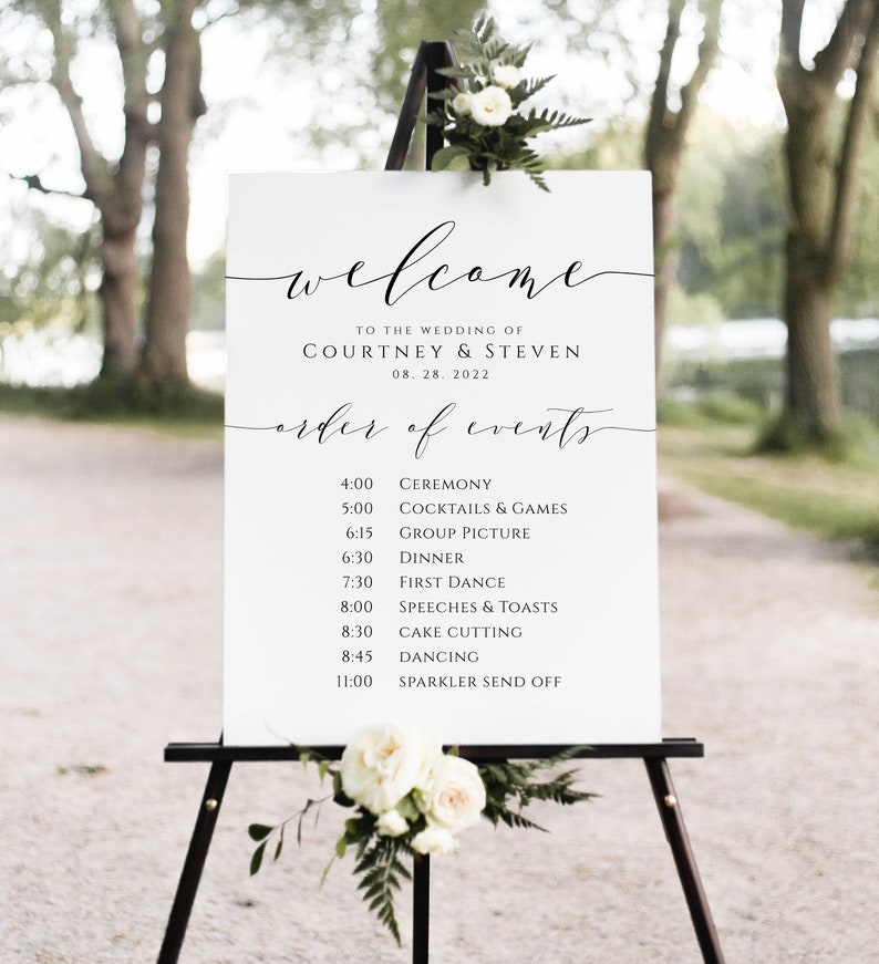 May include: A white welcome sign with black calligraphy for a wedding. The sign lists the order of events for the wedding day, including the ceremony, cocktails and games, group picture, dinner, first dance, speeches and toasts, cake cutting, dancing, and sparkler send off. The sign is on an easel with white flowers.
