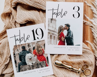 May include: Two white cards with black text and photos. The top card says "Table 3" and has a photo of a couple kissing in front of a building. The bottom card says "Table 19" and has a photo of a couple with a white dog in front of a building. Both cards have the names Charlotte and Anthony and the date 08. 21. 202x.
