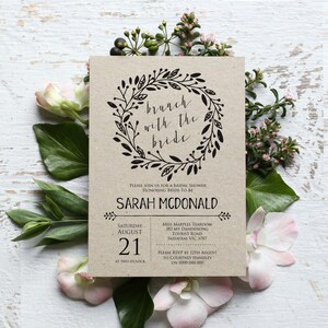May include: A kraft paper bridal shower invitation with a black floral wreath and the text "brunch with the bride". The invitation includes the bride-to-be's name, the date and time of the shower, and the location of the event.
