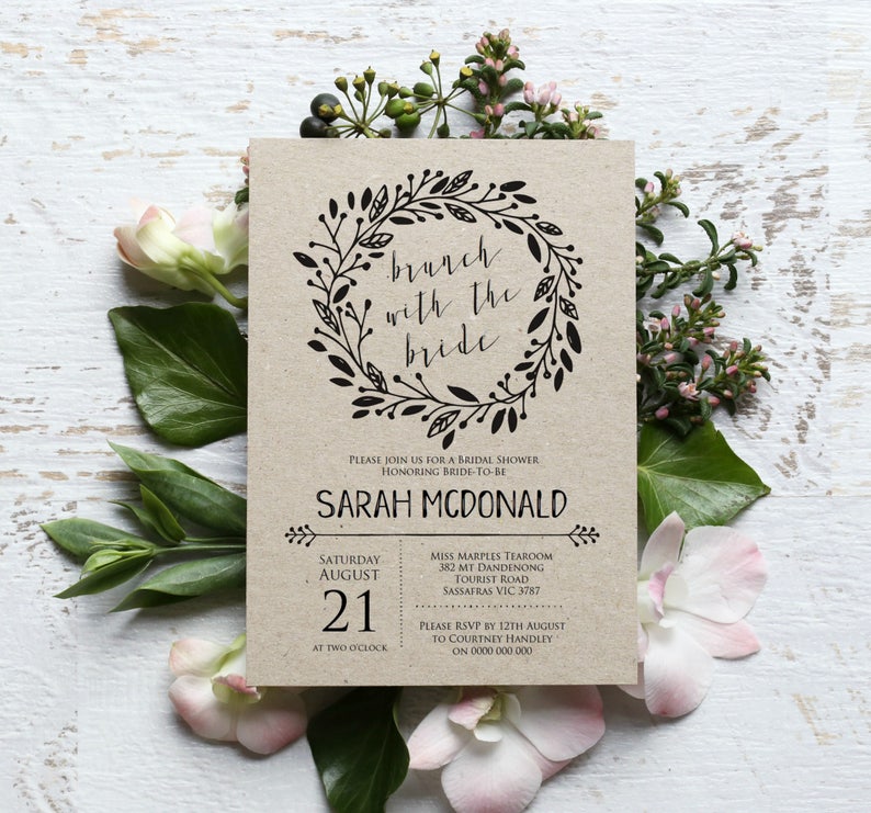 May include: A kraft paper bridal shower invitation with a black floral wreath and the text "brunch with the bride". The invitation includes the bride-to-be's name, the date and time of the shower, and the location of the event.