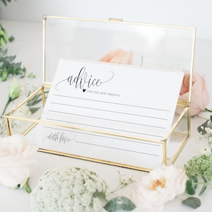 Op de afbeelding: Een gouden glazen doos met een witte kaart erin. De kaart heeft het woord "advice" in cursief geschreven met een hartje en de woorden "for the new parents" eronder. De kaart heeft lijnen om op te schrijven en de woorden "with love" onderaan.