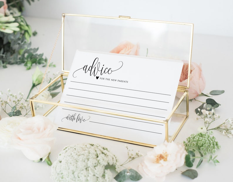 Op de afbeelding: Een gouden glazen doos met een witte kaart erin. De kaart heeft het woord "advice" in cursief geschreven met een hartje en de woorden "for the new parents" eronder. De kaart heeft lijnen om op te schrijven en de woorden "with love" onderaan.