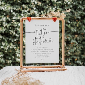 May include: A white sign with black text on a copper pipe stand. The sign reads "Temporary tattoo station" with a heart symbol. Instructions for applying temporary tattoos are listed below.