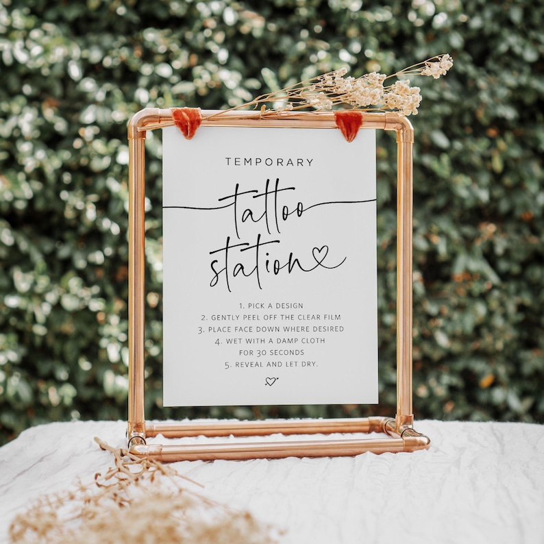 May include: A white sign with black text on a copper pipe stand. The sign reads "Temporary tattoo station" with a heart symbol. Instructions for applying temporary tattoos are listed below.