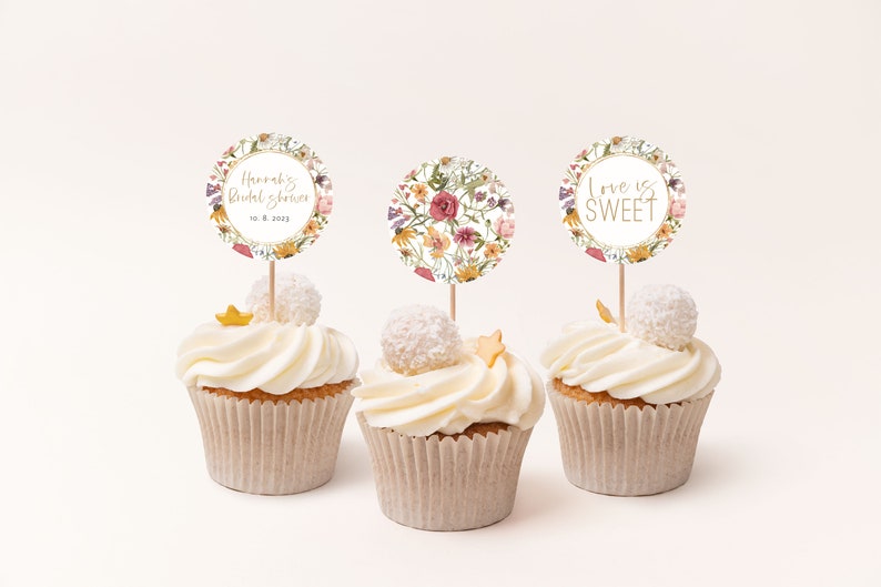 Może przedstawiać: Trzy babeczki z białym lukrem i dekoracyjnymi topperami. Toppery są okrągłe z kwiatowymi wzorami i tekstem &bdquo;Hannah's Bridal Shower 10. 8. 2023&rdquo; i &bdquo;Love is Sweet&rdquo;. Każda babeczka ma element dekoracyjny.