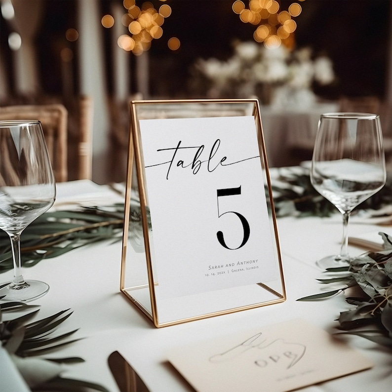 May include: A gold-framed table number card with the text "Table 5" and the names "Sarah and Anthony" with their wedding date and location. The card is on a white table with greenery.