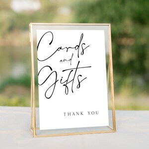 以下が含まれることがあります： 黒い書体で「Cards and Gifts」と書かれた金色の額縁の看板。看板には「Thank You」という黒い文字も書かれている。