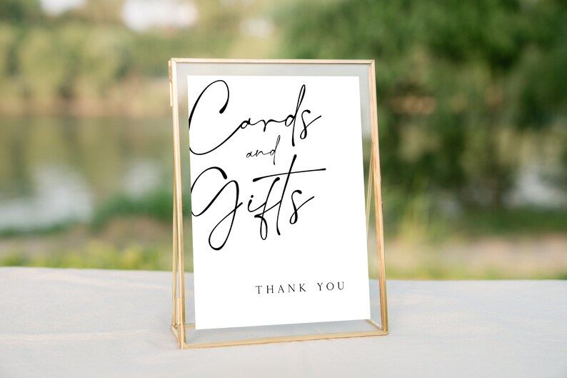 以下が含まれることがあります： 黒い書体で「Cards and Gifts」と書かれた金色の額縁の看板。看板には「Thank You」という黒い文字も書かれている。