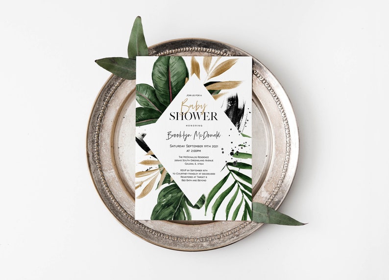 May include: A white baby shower invitation with gold and black accents. The invitation features a diamond shape with the text "Baby Shower" and the name "Brooklyn McDonald". The background features a watercolor design of tropical leaves and gold accents.
