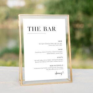 May include: A gold-framed sign with a white background and black text that reads "THE BAR". The sign lists the drinks available at the bar, including beer, wine, spirits, and non-alcoholic options. The sign is on a white table with a blurry background of a lake and trees.
