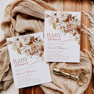 May include: Two baby shower invitations with a floral design. The invitations are on a wooden surface with a beige fabric and gold wax seals. The text on the invitations reads "Baby Shower" and "Join us for a baby shower honoring Hannah Thompson". The date and time of the shower are listed as "Sat | Nov 17 | 2PM". The location is listed as "The Smith Residence, 276 Willow Lane, Galena, IL". The RSVP information is listed as "Kindly RSVP Courtney 000.000.0000". The invitations also include the text "Registered at Target & Amazon.com".