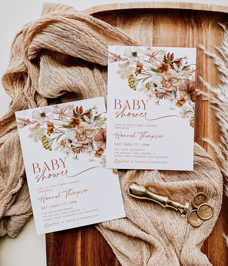 May include: Two baby shower invitations with a floral design. The invitations are on a wooden surface with a beige fabric and gold wax seals. The text on the invitations reads "Baby Shower" and "Join us for a baby shower honoring Hannah Thompson". The date and time of the shower are listed as "Sat | Nov 17 | 2PM". The location is listed as "The Smith Residence, 276 Willow Lane, Galena, IL". The RSVP information is listed as "Kindly RSVP Courtney 000.000.0000". The invitations also include the text "Registered at Target & Amazon.com".