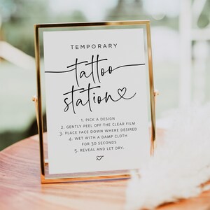 May include: A gold-framed sign with the text "TEMPORARY tattoo station" in black script. Instructions for applying temporary tattoos are listed below: 1. Pick a design, 2. Gently peel off the clear film, 3. Place face down where desired, 4. Wet with a damp cloth for 30 seconds, 5. Reveal and let dry.