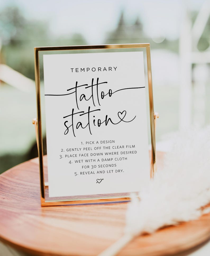 May include: A gold-framed sign with the text "TEMPORARY tattoo station" in black script. Instructions for applying temporary tattoos are listed below: 1. Pick a design, 2. Gently peel off the clear film, 3. Place face down where desired, 4. Wet with a damp cloth for 30 seconds, 5. Reveal and let dry.