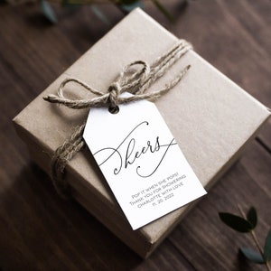 Könnte beinhalten: Ein weißes Geschenkanhänger mit schwarzer Kalligraphie, auf dem "Cheers" und der Text "Pop it when she pops! Thank you for showering Charlotte with love 11.20.2022" steht. Der Anhänger ist mit einer Schnur an einer braunen Geschenkbox gebunden.