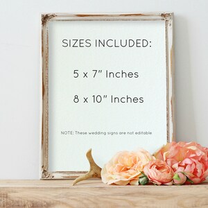 May include: A white frame with a white background and black text that reads "SIZES INCLUDED: 5 x 7" Inches 8 x 10" Inches NOTE: These wedding signs are not editable". The frame is sitting on a wooden surface with a white antler and peach-colored flowers.