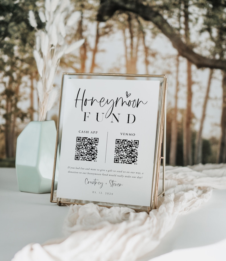 May include: A white sign with a gold frame, announcing a honeymoon fund. The sign has a black heart symbol and the text "Honeymoon Fund". It also includes QR codes for Cash App and Venmo, with the text "Cash App" and "Venmo" above each code. The sign also includes the text "If you had fun and want to give a gift to send us on our way, a donation to our honeymoon fund would really make our day!" and the names "Courtrey & Stever" with the date "05.12.202x".