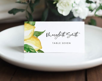 May include: A white place card with a watercolor illustration of lemons and green leaves. The text on the card reads "Marybeth Smith" and "Table Seven".