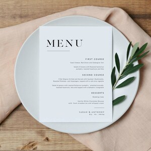 May include: A wedding menu card with a light blue background and black text. The menu includes a first course, second course, and desserts. The card is on a white plate with a green olive branch.