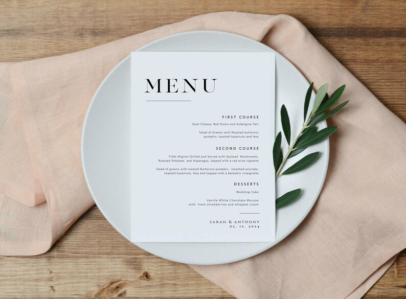 May include: A wedding menu card with a light blue background and black text. The menu includes a first course, second course, and desserts. The card is on a white plate with a green olive branch.