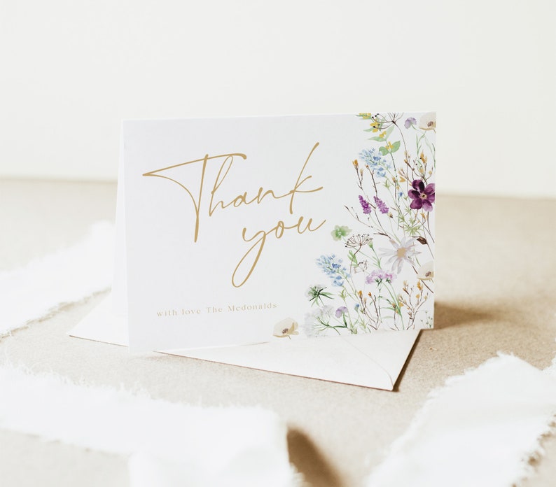 May include: A white thank you card with gold lettering that says "Thank you" and "with love, The McDonalds". The card has a floral design on the right side with purple, blue, and white flowers.